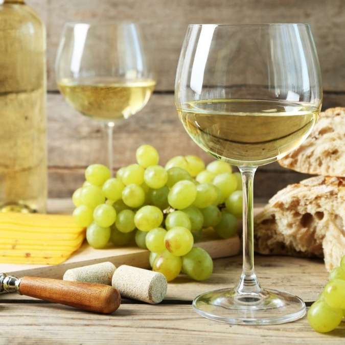 A glass of white wine in the foreground with a second glass and a bottle behind it, arranged on a rustic wooden table with green grapes, bread, a cheese knife, and corks.