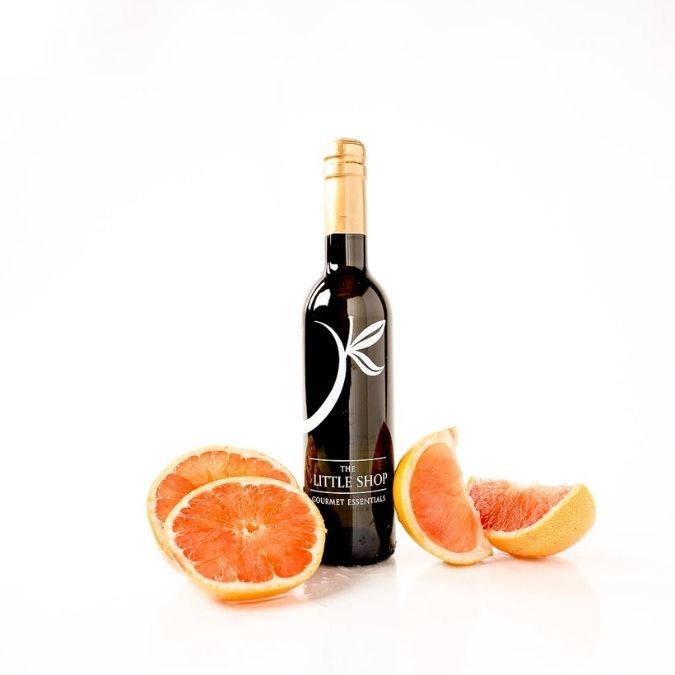 A bottle of grapefruit white balsamic vinegar from 'The Little Shop' stands on a white reflective surface, flanked by fresh grapefruit slices showing their pink-orange pulp.