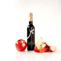 A bottle of balsamic vinegar by The Little Shop, styled with a whole red apple and apple slices on a white reflective surface.