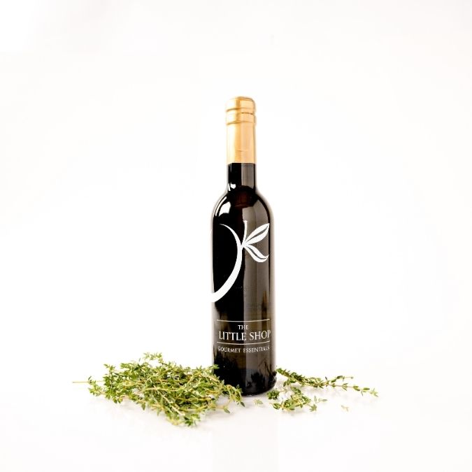 A dark glass bottle of Herbes de Provence Olive Oil by 'The Little Shop' with a gold foil cap and white logo, styled with fresh green thyme sprigs on a clean white background.