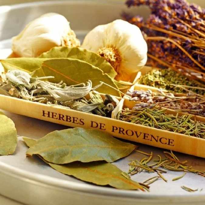 A wooden box labeled 'HERBES DE PROVENCE' filled with dried herbs, bay leaves, garlic bulbs, and lavender sprigs on a white plate.