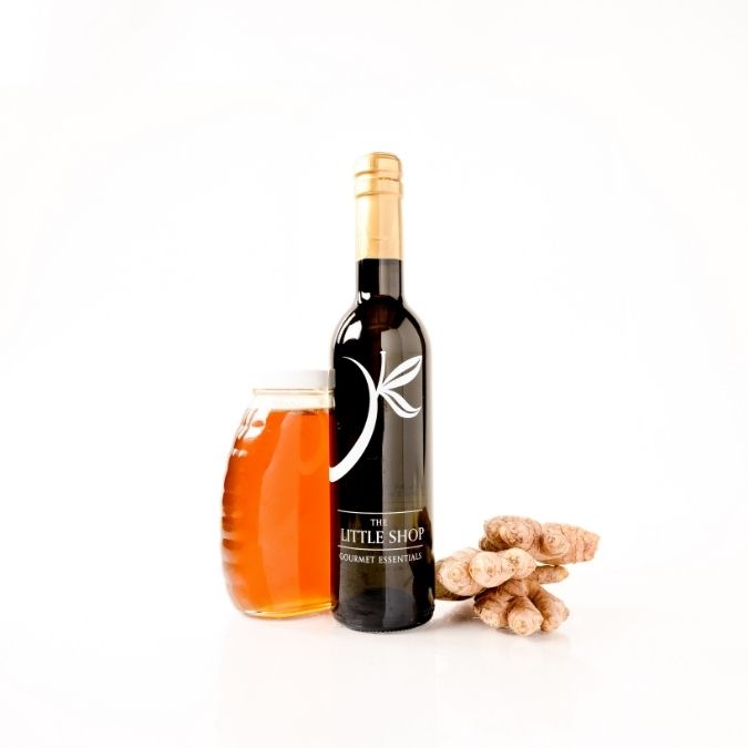 A bottle of gourmet honey ginger white balsamic next to a glass jar of honey with ginger root pieces on a clean white background. The bottle label reads 'THE LITTLE SHOP GOURMET ESSENTIALS'.