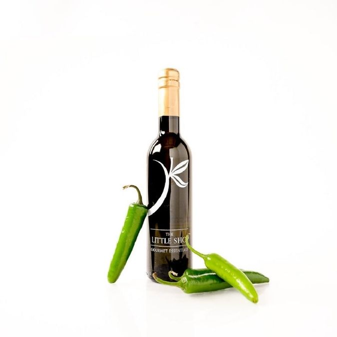 A bottle of Jalapeno Olive Oil stands on a white background, styled with three fresh green jalapeno peppers.