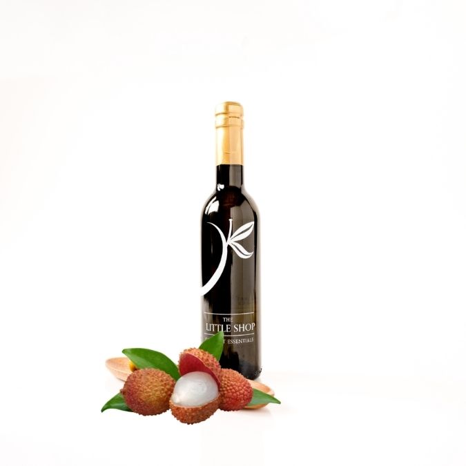 A bottle of gourmet white balsamic vinegar from 'THE LITTLE SHOP' on a clean white background with whole lychee fruit styled around it. 