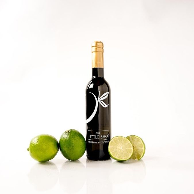 A bottle of balsamic vinegar with a black label and gold foil cap stands on a white reflective surface, surrounded by whole and sliced limes on both sides.