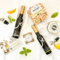 A styled flat-lay of gourmet food items on a white textured fabric background, including two dark glass bottles with gold caps, a packaged pasta dish, a jar of artichoke, a small bag of herb blend, and lemon slices and basil.