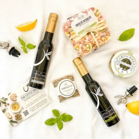 A styled flat-lay of gourmet food items on a white textured fabric background, including two dark glass bottles with gold caps, a packaged pasta dish, a jar of artichoke, a small bag of herb blend, and lemon slices and basil.