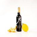 A bottle of wine with a gold foil top and a black label featuring a white stylized leaf logo, styled with fresh yellow lemon slices and a whole lemon on a clean white background. The label reads 'THE LITTLE SHOP'.