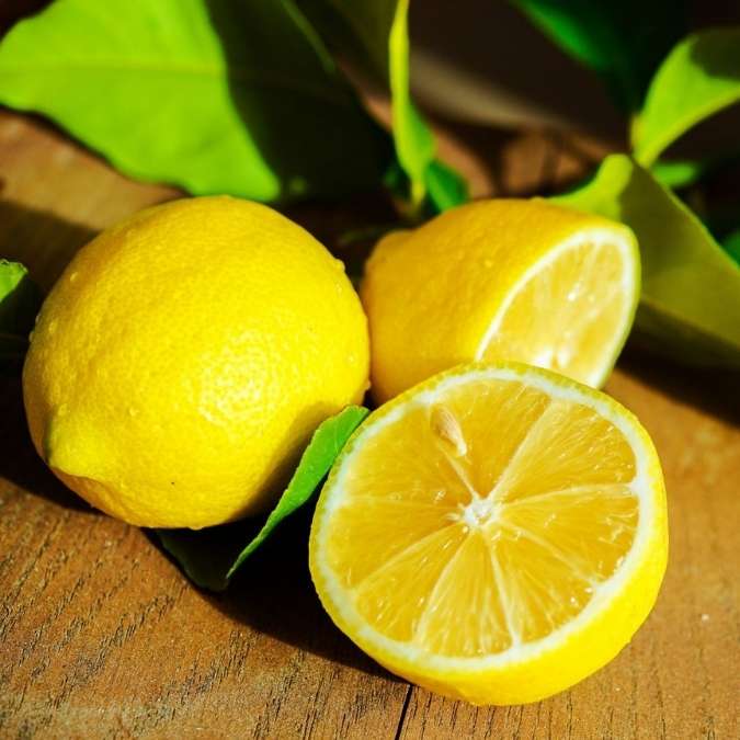Three lemons on a wooden surface with green leaves in the background, including one whole lemon, one cut in half showing the juicy interior, and one partially visible behind.