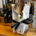 Gift set with wooden spoons, white napkins, and black ribbon on a wooden table with olive oil bottles in the background.