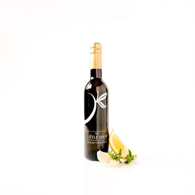 Dark glass bottle of Milanese Gremolata Olive Oil on a white background with a slice of fresh lemon, garlic cloves, and green herbs. 