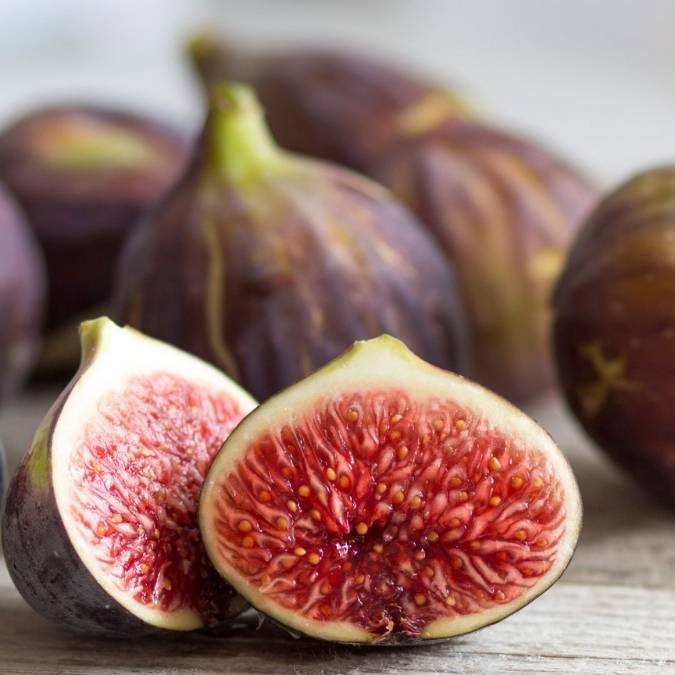 Fresh figs on a rustic wooden surface, with two halves cut open in the foreground showing pink-red interiors and purple skins, with whole figs blurred in the background.