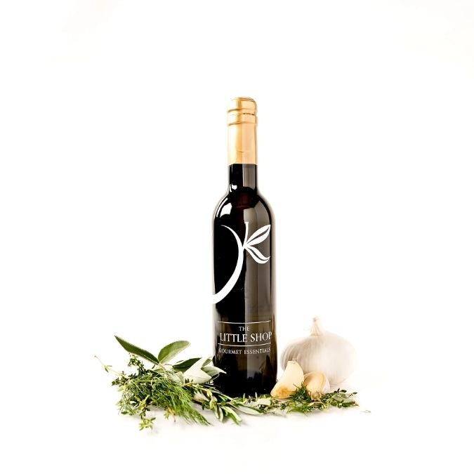 A bottle of gourmet balsamic vinegar from 'The Little Shop' is displayed on a clean white background, styled with fresh herbs, garlic, and a whole garlic bulb.