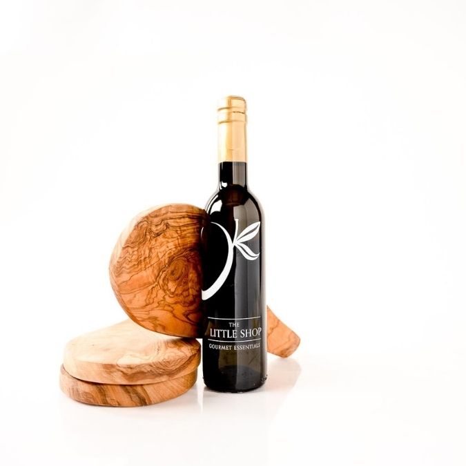 A dark glass bottle of gourmet olive oil with a gold foil top and white logo, styled with olive wood coasters and a wooden box on a clean white background.