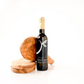 A dark glass bottle of gourmet olive oil with a gold foil top and white logo, styled with olive wood coasters and a wooden box on a clean white background.