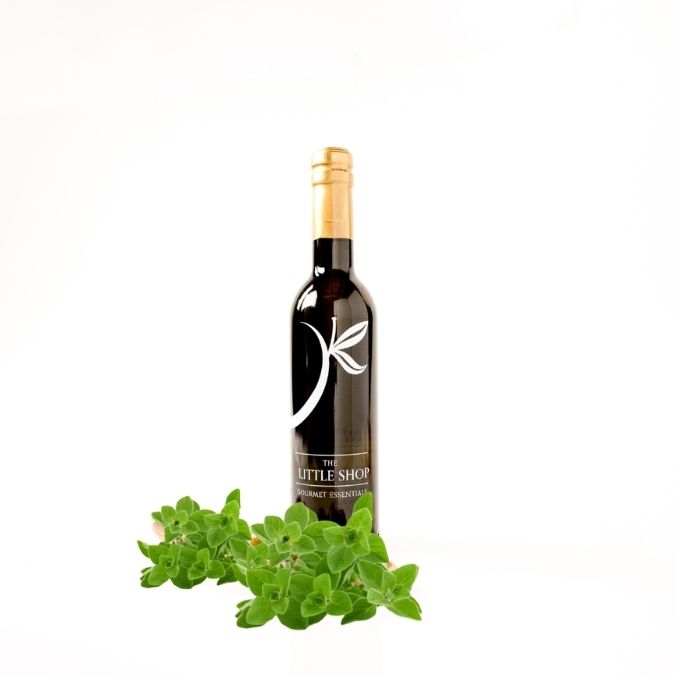 A glass bottle of gourmet white balsamic vinegar from 'THE LITTLE SHOP' on a white background with fresh oregano leaves styled around the bottle. 