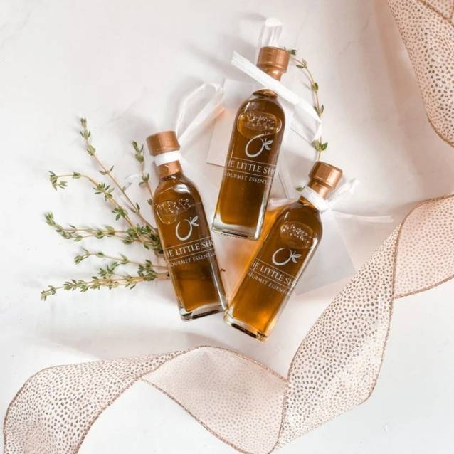 Three small glass bottles of olive oil arranged on a white surface with decorative sprigs and a sheer beige ribbon. The labels read 'Little Sister' and 'Extra Virgin Olive Oil.