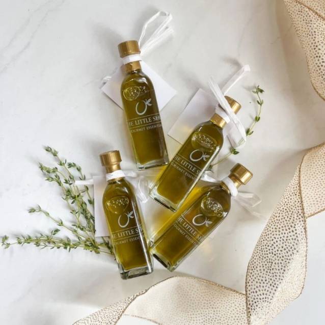 Four small glass bottles of olive oil with gold caps and white ribbons, arranged on a light marble surface with decorative herbs and beige fabric.