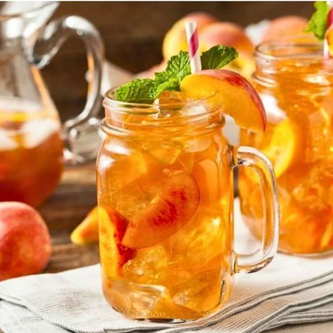 Glass mug with peach iced tea garnished with peach slices and mint leaves on a wooden surface.