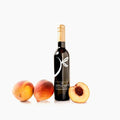 A bottle of gourmet peach white balsamic vinegar from 'The Little Shop' stands upright on a white background, flanked by two whole peaches and one halved peach on the ground level.