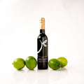 A bottle of Persian Lime Olive Oil with a  gold foil cap stands upright on a white reflective surface, surrounded by five green limes on both sides. The label reads 'THE LITTLE SHOP'.