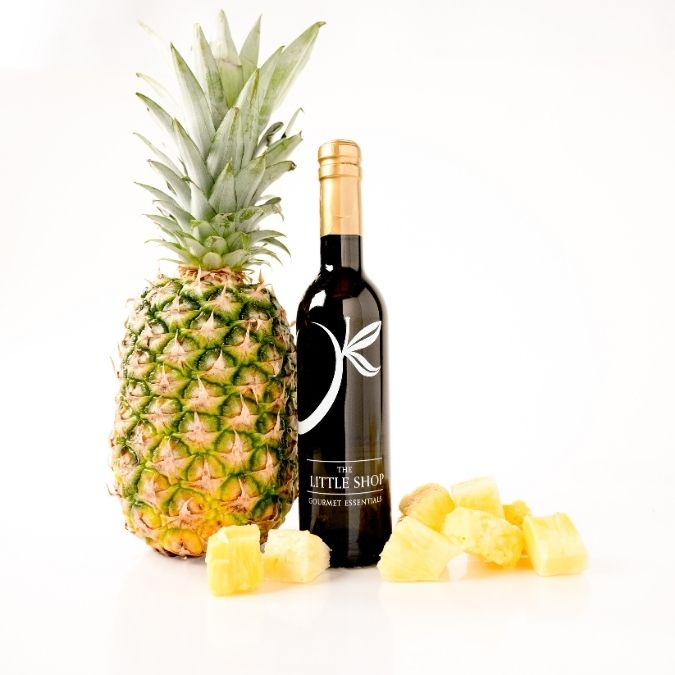 A bottle of gourmet balsamic vinegar by The Little Shop, styled with a whole pineapple, a few pineapple rinds, and several pineapple chunks on a clean white background.