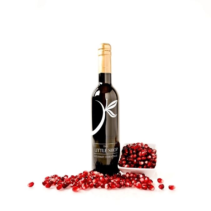A bottle of gourmet balsamic vinegar, styled with scattered red pomegranate seeds and a small bowl of pomegranates on a clean white background.