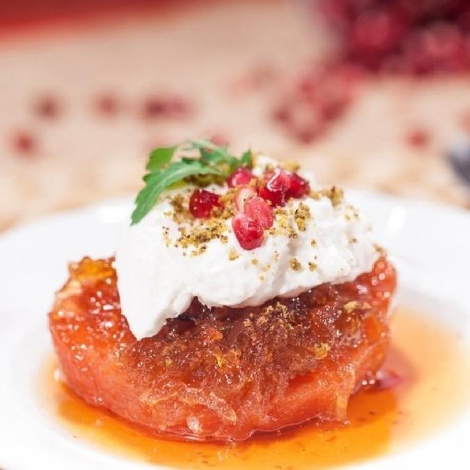 A close-up of a dessert on a white plate, featuring a baked orange-red cake or bread topped with whipped cream, pomegranate seeds, a green herb garnish, and a sprinkle of chopped nuts, with balsamic glaze pooling around the base.