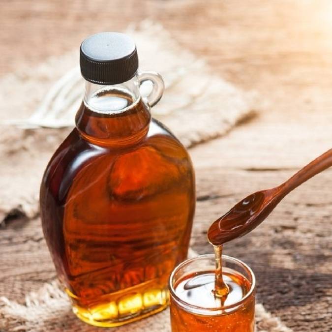 A clear glass bottle filled with maple syrup, shown beside a small glass and a wooden spoon dripping the syrup onto a rustic wooden surface.