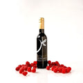 A dark bottle with balsamic vinegar, a gold foil top and a white stylized leaf logo on the front, standing on a white reflective surface with red raspberries scattered around its base. The label reads 'THE LITTLE SHOP'.