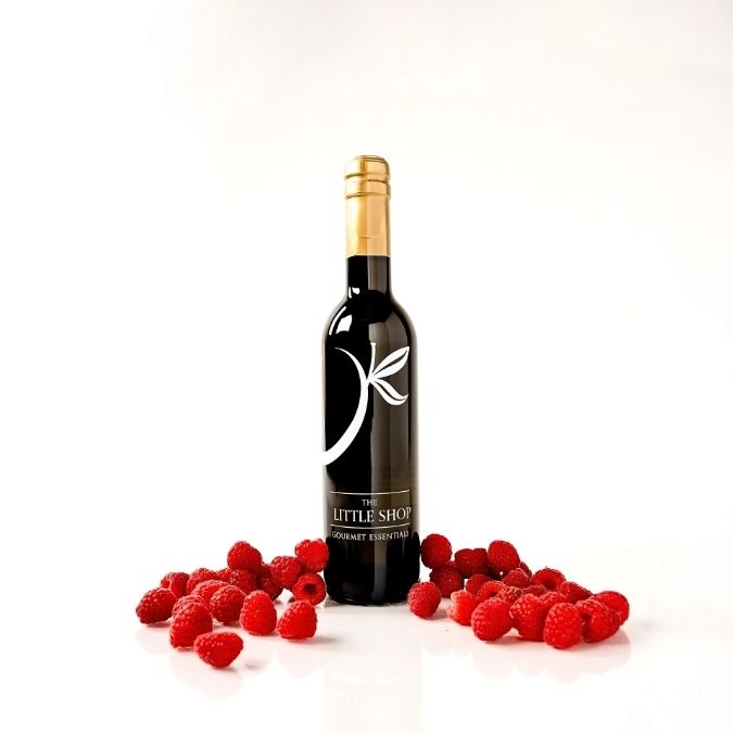 A dark bottle with balsamic vinegar, a gold foil top and a white stylized leaf logo on the front, standing on a white reflective surface with red raspberries scattered around its base. The label reads 'THE LITTLE SHOP'.