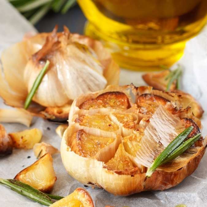 Roasted garlic bulbs with crispy browned edges, served on parchment paper with rosemary sprigs, accompanied by a glass container of olive oil in the background.