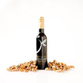 A bottle of gourmet roasted walnut oil stands upright on a white background, flanked by scattered pieces of walnuts. The bottle is dark glass with a gold foil top and a white logo reading 'THE LITTLE SHOP'.