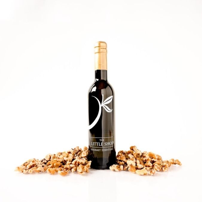 A bottle of gourmet roasted walnut oil stands upright on a white background, flanked by scattered pieces of walnuts. The bottle is dark glass with a gold foil top and a white logo reading 'THE LITTLE SHOP'.