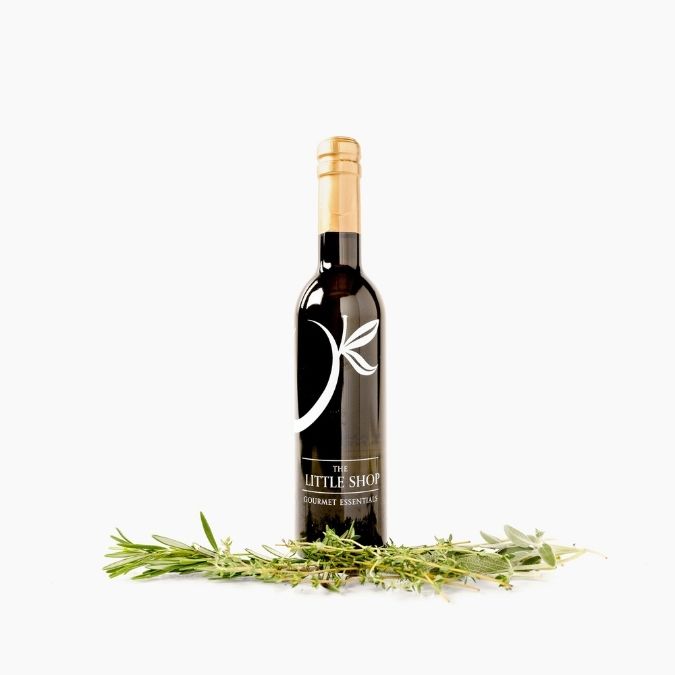 A bottle of gourmet rosemary olive oil with a black label and gold foil cap, styled with fresh rosemary sprigs on a clean white background. The label features a white stylized leaf logo and the brand name 'The Little Shop'.