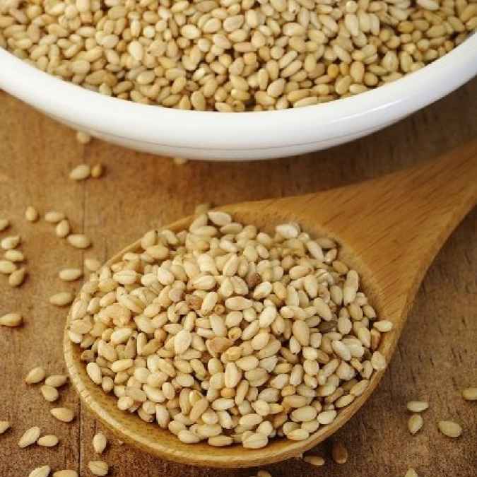Raw sesame seeds in a wooden spoon with a white bowl of sesame seeds in the background on a wooden surface.