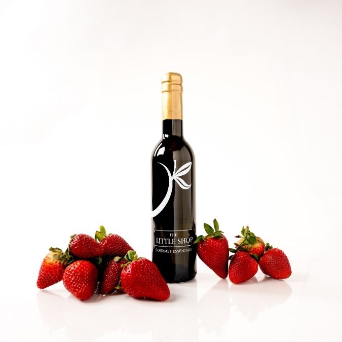 A bottle of balsamic vinegar with a black label and gold foil cap stands on a white reflective surface, surrounded by several fresh red strawberries on both sides. 