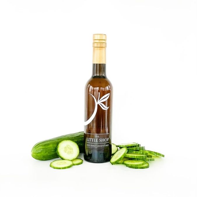 A bottle of gourmet balsamic vinegar by The Little Shop, styled with fresh whole and sliced cucumbers on a clean white background.