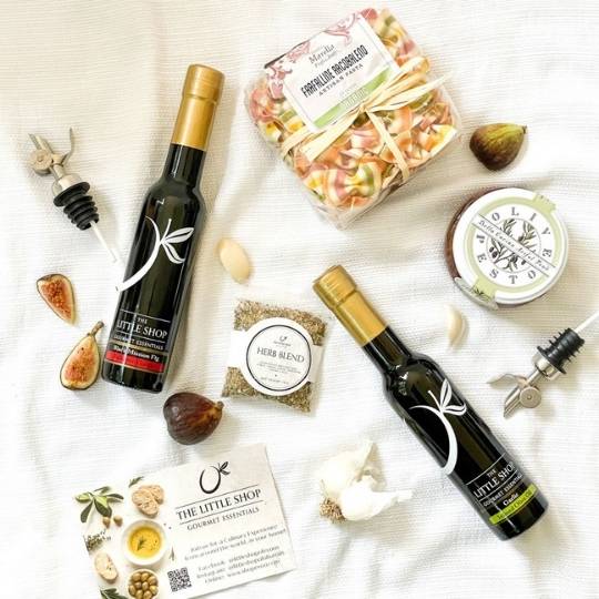 A styled flat-lay of gourmet food items from 'The Little Shop' on a white textured fabric background. The assortment includes two bottles of coconut oil with gold caps, a bag of fig and coconut flakes, a jar of coconut essential oil, a herb blend packet, garlic cloves, figs, and a bottle stopper.