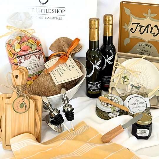 A gourmet gift basket arrangement featuring a bottle of olive oil, a bottle of balsamic vinegar, a jar of herb blend, a small knife, a wooden cutting board, oil bottles, a burlap-wrapped item, a bag of pasta, and a white cloth on a white background.