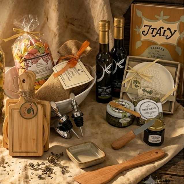 A gourmet gift hamper featuring olive oil, honey, soap, and wooden kitchen accessories arranged on a rustic beige fabric backdrop. The set includes bottles of olive oil, a box of Italy recipe cookies, a jar of honey, a soap bar, a wooden cutting board, and a wooden spoon.