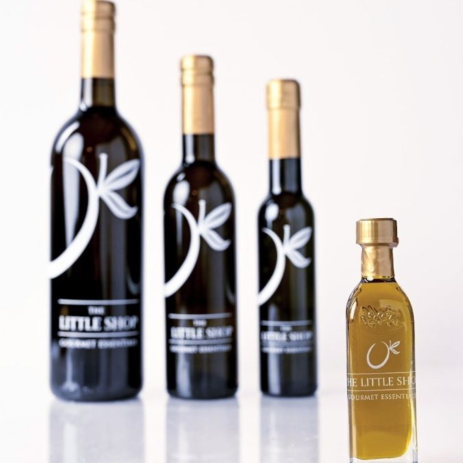 Four dark glass bottles with gold caps and white labels reading 'The Little Shop Gourmet Essential' are arranged on a white reflective surface against a white background, with the front bottle in sharp focus and the others softly blurred.