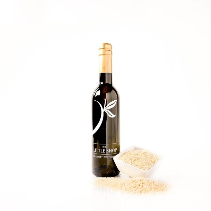 A dark glass bottle of gourmet toasted sesame oil on a reflective white surface styled with a small square bowl of sesame seeds and scattered sesame seeds around the bottle. 
