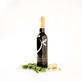A dark glass bottle of wine with a gold foil cap and a white logo, standing upright on a white background with sprigs of rosemary and garlic cloves placed at its base. The label reads 'The Little Shop' and 'Grenache Prosecco'.