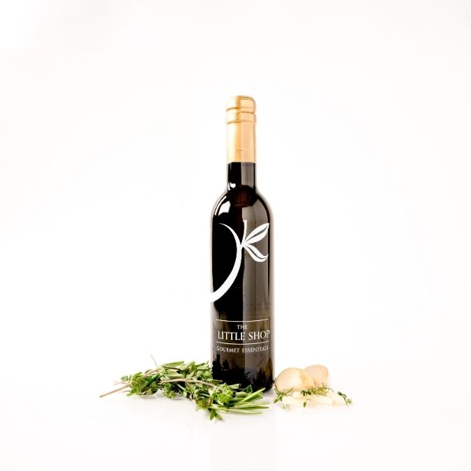 A dark glass bottle of wine with a gold foil cap and a white logo, standing upright on a white background with sprigs of rosemary and garlic cloves placed at its base. The label reads 'The Little Shop' and 'Grenache Prosecco'.