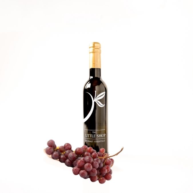 A bottle ultra white balsamic vinegar from 'The Little Shop' with a gold foil top and a white leaf logo, displayed on a white background with a bunch of red grapes at its base.