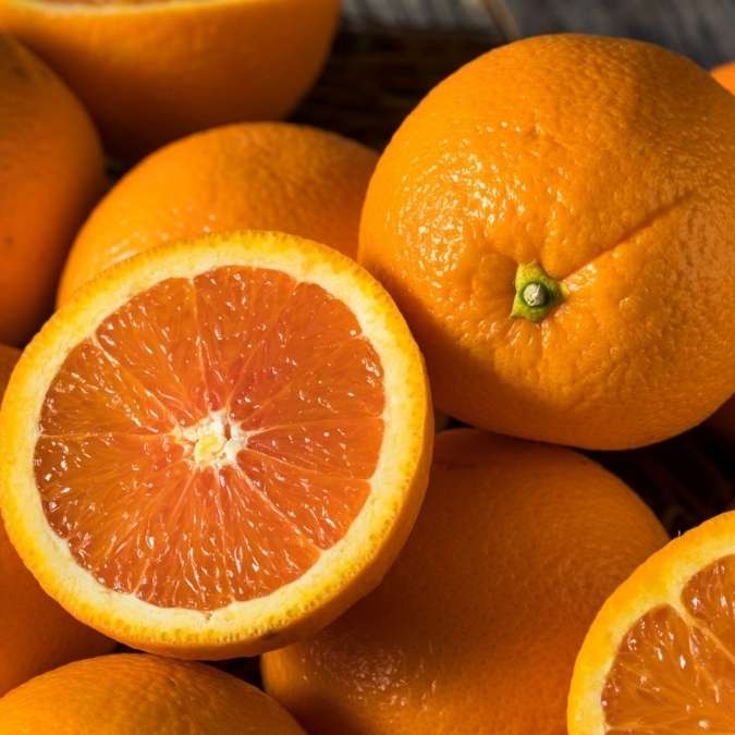 A close-up pile of fresh oranges, including one whole orange and one cut in half to show the juicy segmented pulp.
