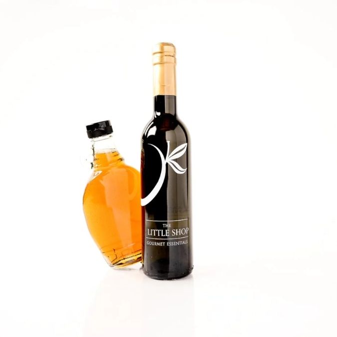 Two bottles from 'The Little Shop Gourmet Essentials' on a white background: a dark glass bottle with a gold foil top and white logo, and a clear glass bottle filled with Vermont maple syrup and a black cap.