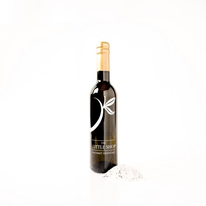 Dark glass bottle of white truffle premium olive oil labeled with 'THE LITTLE SHOP' on a crisp white background with shaved white truffles surrounding the bottle. 