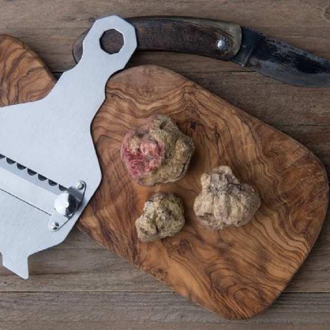 A metal food prep tool with a serrated edge rests on a wooden board alongside three raw truffles, with a knife in the background on a rustic wood surface.
